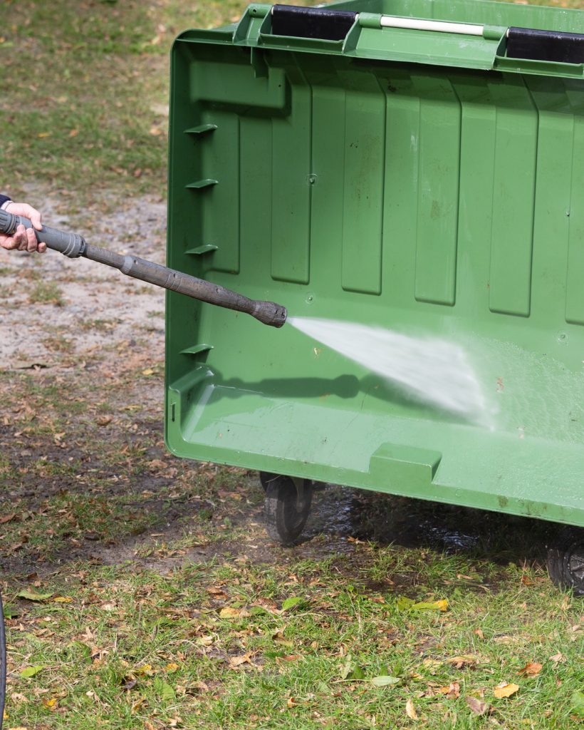 A municipal service worker treats a garbage container with a disinfectant solution. Washing the garbage container.